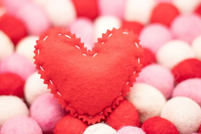 A stitched red felt heart resting on a bed of pink, red, and white pom-poms